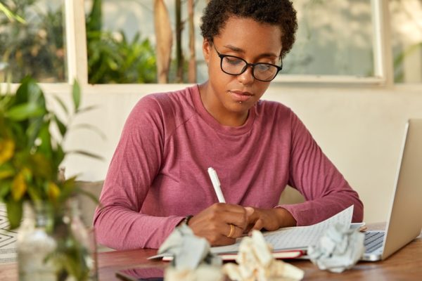 Black lady creats publication, writes records in notepad, focused on writing, uses laptop computer for searching information in internet, sits at work place with crumpled paper creats test examination
