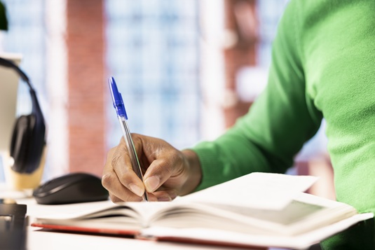 Man reading and writing for school exam on notepad at home office desk, close up. African american student doing homework, writing down info with pen, researching for university